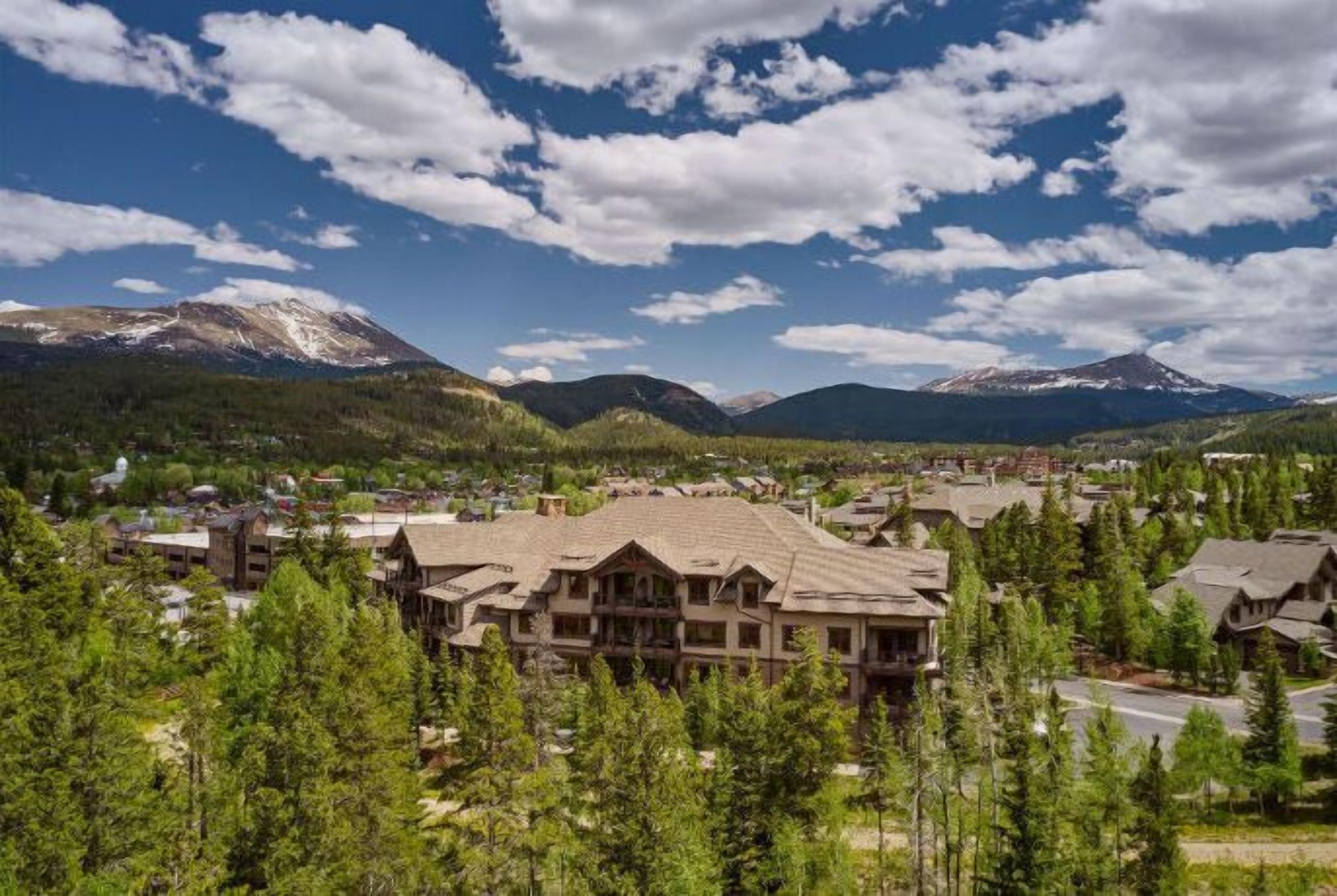 A lodge with a large central building surrounded by trees, set against a backdrop of mountains and a clear sky.