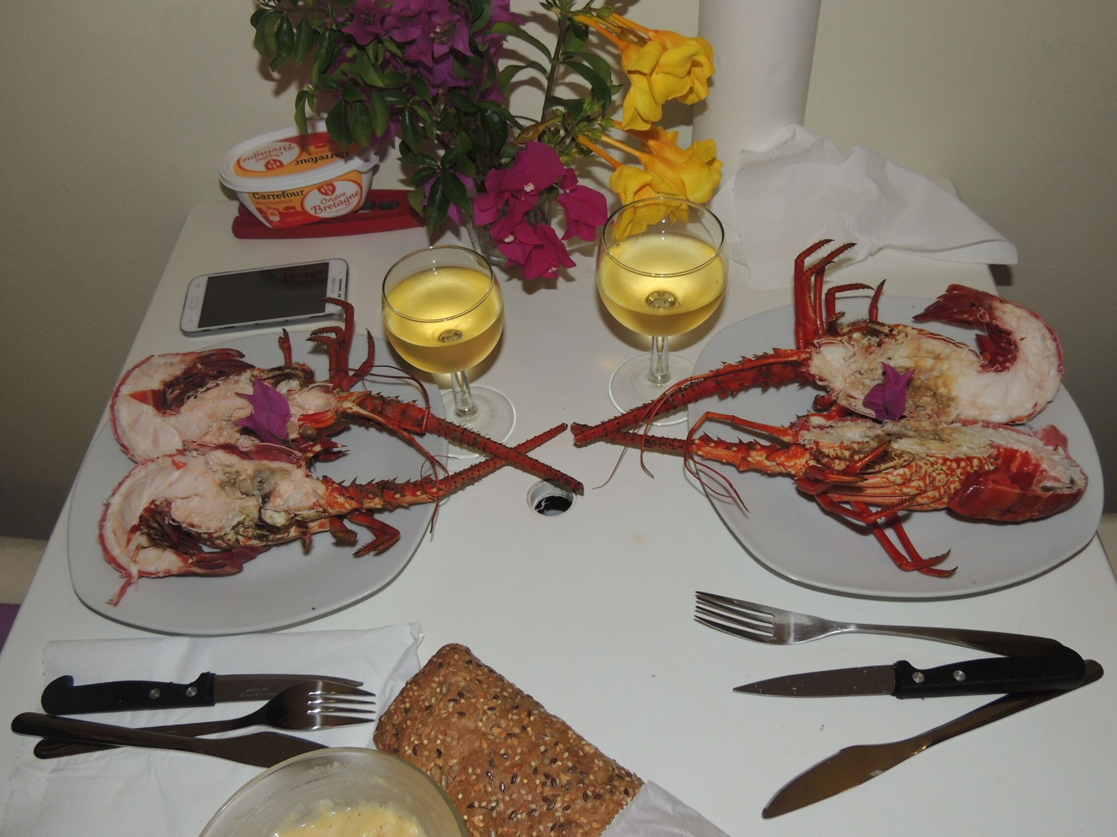 A dining table with two plates of cooked lobsters, a glass of white wine, a glass of red wine, a loaf of bread, and a bowl of butter.