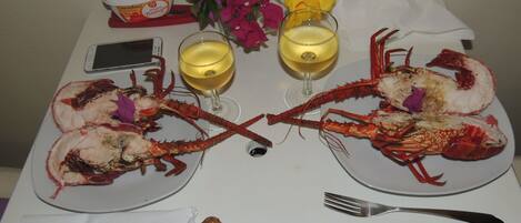 A dining table with two plates of cooked lobsters, a glass of white wine, a glass of red wine, a loaf of bread, and a bowl of butter.