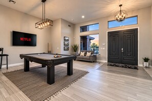 Un salon moderne avec une table de billard, un canapé, un téléviseur diffusant du contenu « NETFLIX » et un lustre.