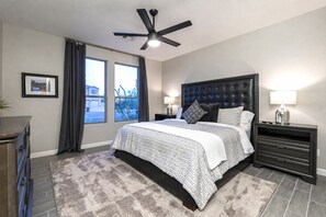 Une chambre à coucher comprenant un lit, un ventilateur de plafond, une table de chevet avec une lampe, une commode et une fenêtre avec des rideaux.