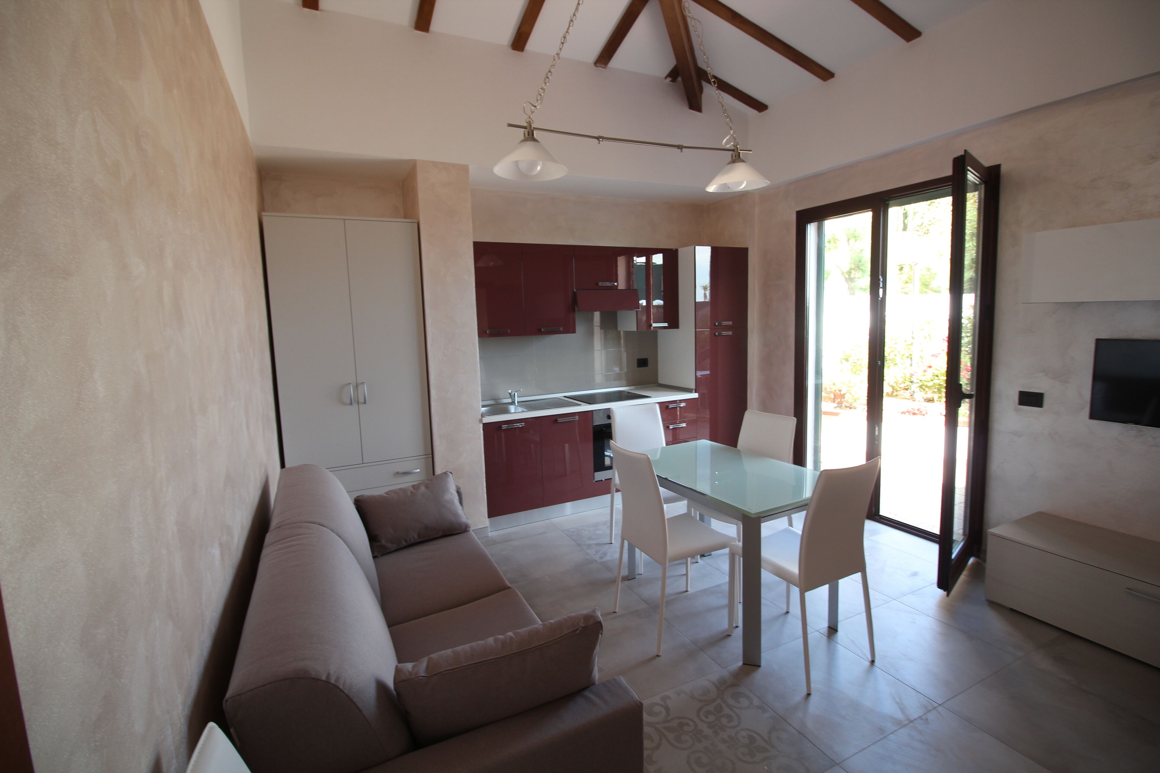 A modern living room with a sofa, dining table, and chairs. There is a kitchen area with cabinets and a sink. The room has a large window and a flat-screen TV mounted on the wall.