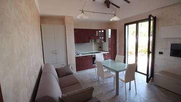 A modern living room with a sofa, dining table, and chairs. There is a kitchen area with cabinets and a sink. The room has a large window and a flat-screen TV mounted on the wall.