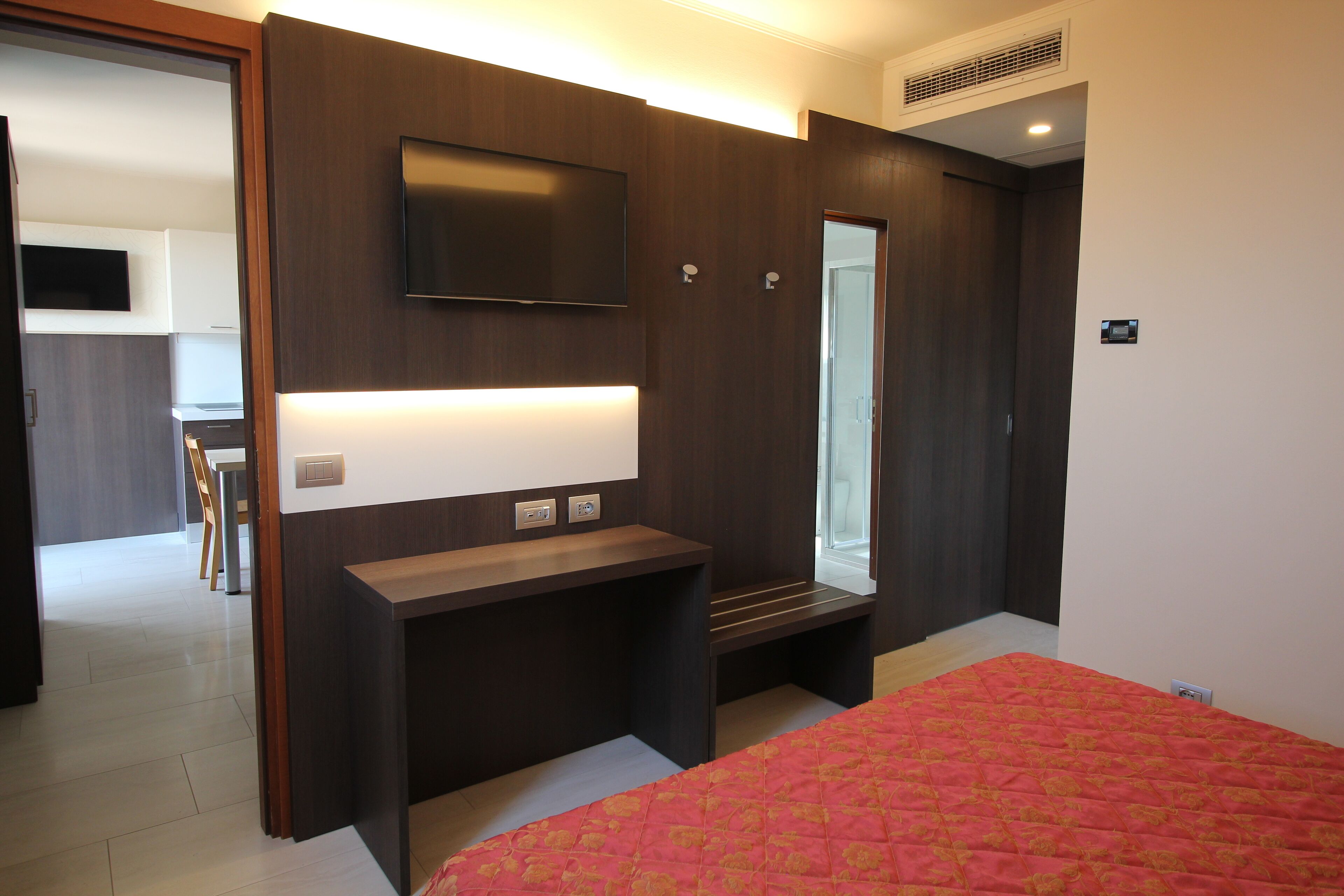 A modern bedroom with a large bed, a bedside table, a mirror, and a TV mounted on the wall.
