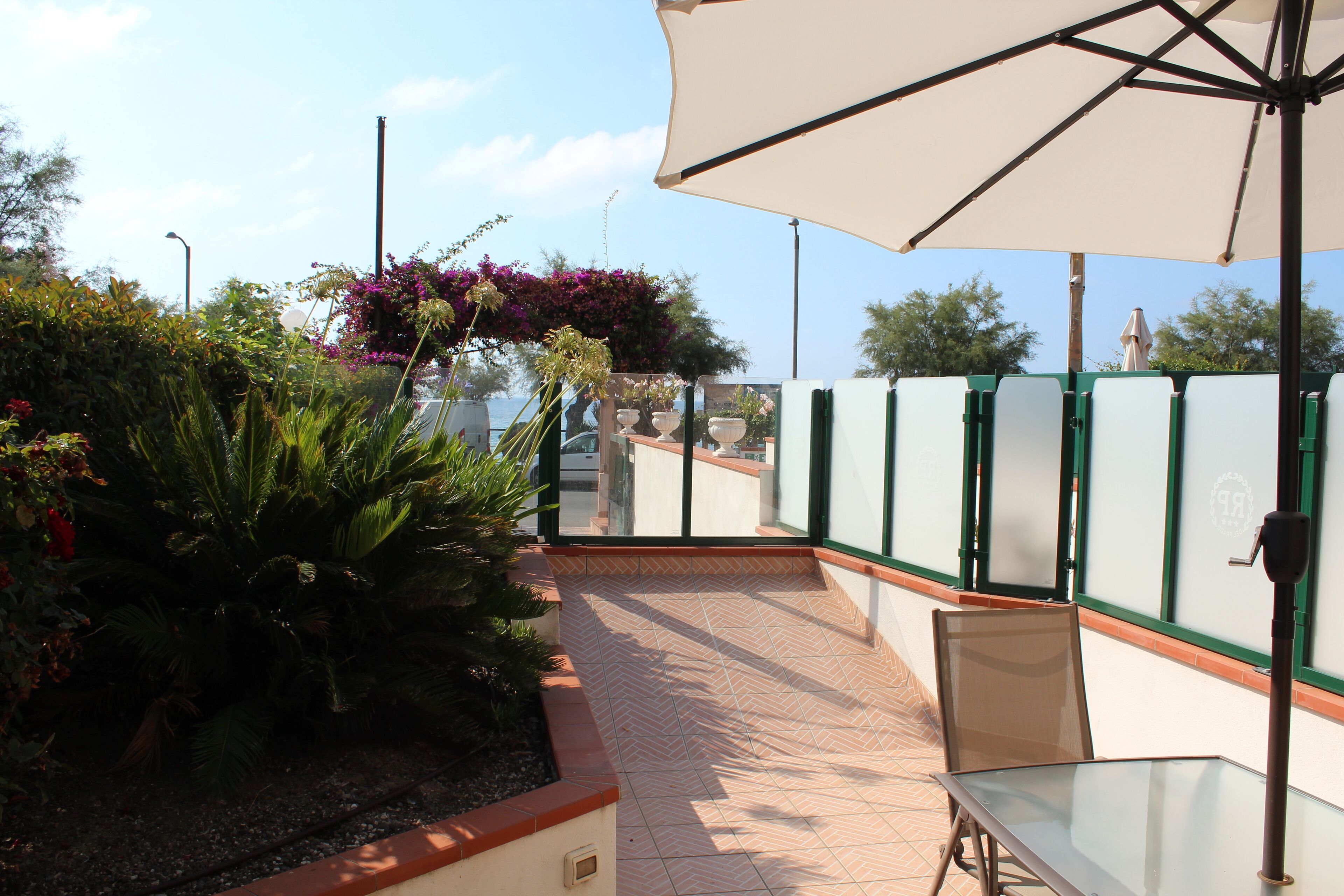 A patio area with a glass table and chairs, surrounded by plants and a large umbrella.