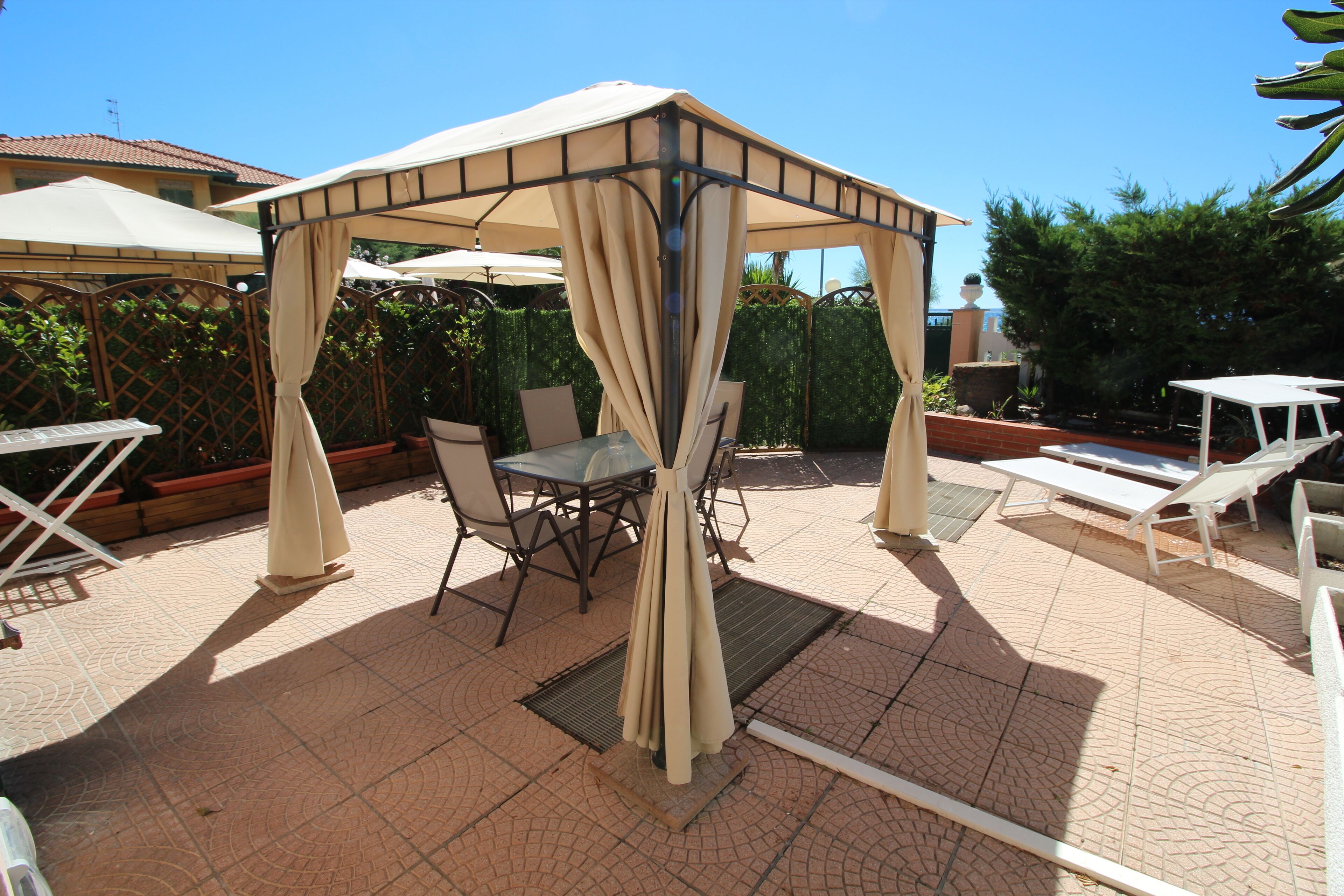 A covered outdoor seating area with tables and chairs, a pergola structure, and a pool in the background.
