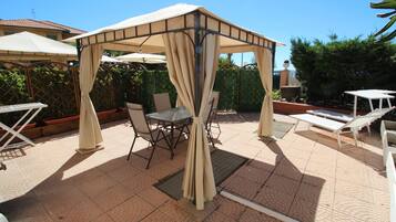 A covered outdoor seating area with tables and chairs, a pergola structure, and a pool in the background.