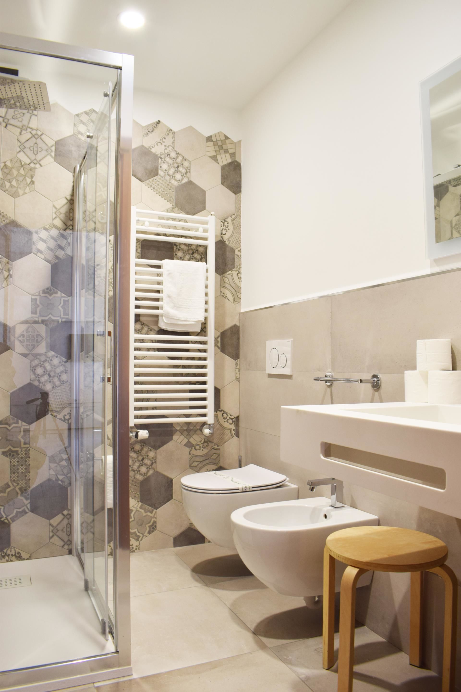 A modern bathroom with a glass shower enclosure, a toilet, a bidet, and a wooden stool.