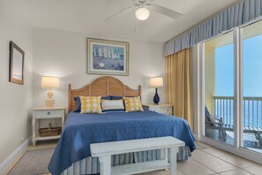 Ein Schlafzimmer mit einem großen Bett, zwei Nachttischen, einem Deckenventilator, einem Fenster mit Vorhängen, einem Balkon und einem gerahmten Bild an der Wand.