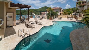 A swimming pool with lounge chairs and a covered area, surrounded by buildings and a mountainous backdrop. - Grande Bay Resort Condo - Come Stay at 'Sunsets and Breezes' (Cruz Bay)