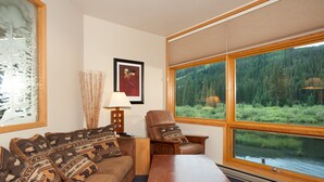 Ein gemütliches Wohnzimmer mit großem Fenster, einer Couch mit gemusterten Kissen, einem Holztisch und einem gerahmten Bild an der Wand.