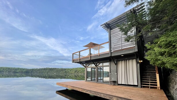A modern house with a wooden deck extending over a lake, large glass doors, and a balcony with a pergola. - Meech Lake peaceful retreat (Chelsea)