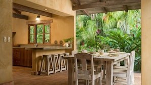 A rustic kitchen with a wooden table and chairs, a window with a view of tropical plants, and a bar area with stools. - Private,Secluded Tamarindo Beach House -DIRECT BEACH ACCESS Perfect for families (Playa Tamarindo)