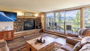 A cozy living room with a stone fireplace, a large TV, and a balcony with a mountain view. - Mountain & Golf Course Views, Close to Shuttle Stop for Easy Resort Access! (Keystone)