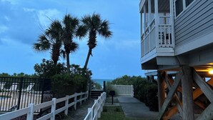 Une propriété située en bord de mer, dotée d’une terrasse en bois, d’une clôture blanche et d’un palmier.
