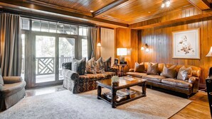 Un salón acogedor con techo de madera, un ventanal con cortinas, un sofá de cuero marrón, un sillón estampado y una mesa de centro con una planta.