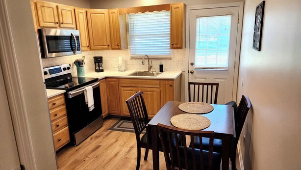 A compact kitchen with wooden cabinets, a stainless steel microwave, a built-in oven, and a coffee maker. There's a dining table with two chairs and a window with blinds.