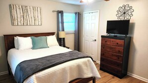 A bedroom with a bed, a dresser, a TV, a window with curtains, and a wall art piece.