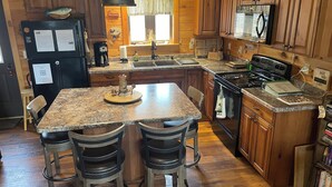 A kitchen with wooden cabinets, a granite countertop, a stove, a sink, and bar stools. - Upscale Cozy Cabin in the Woods, LETCHWORTH, Stony Brook Parks, Wineries (Dalton)
