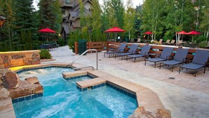 A hot tub surrounded by lounge chairs and red umbrellas, with a stone wall and a view of trees and buildings in the background. - Huge Mountain Condo with Fireplace & Balcony (Edwards)