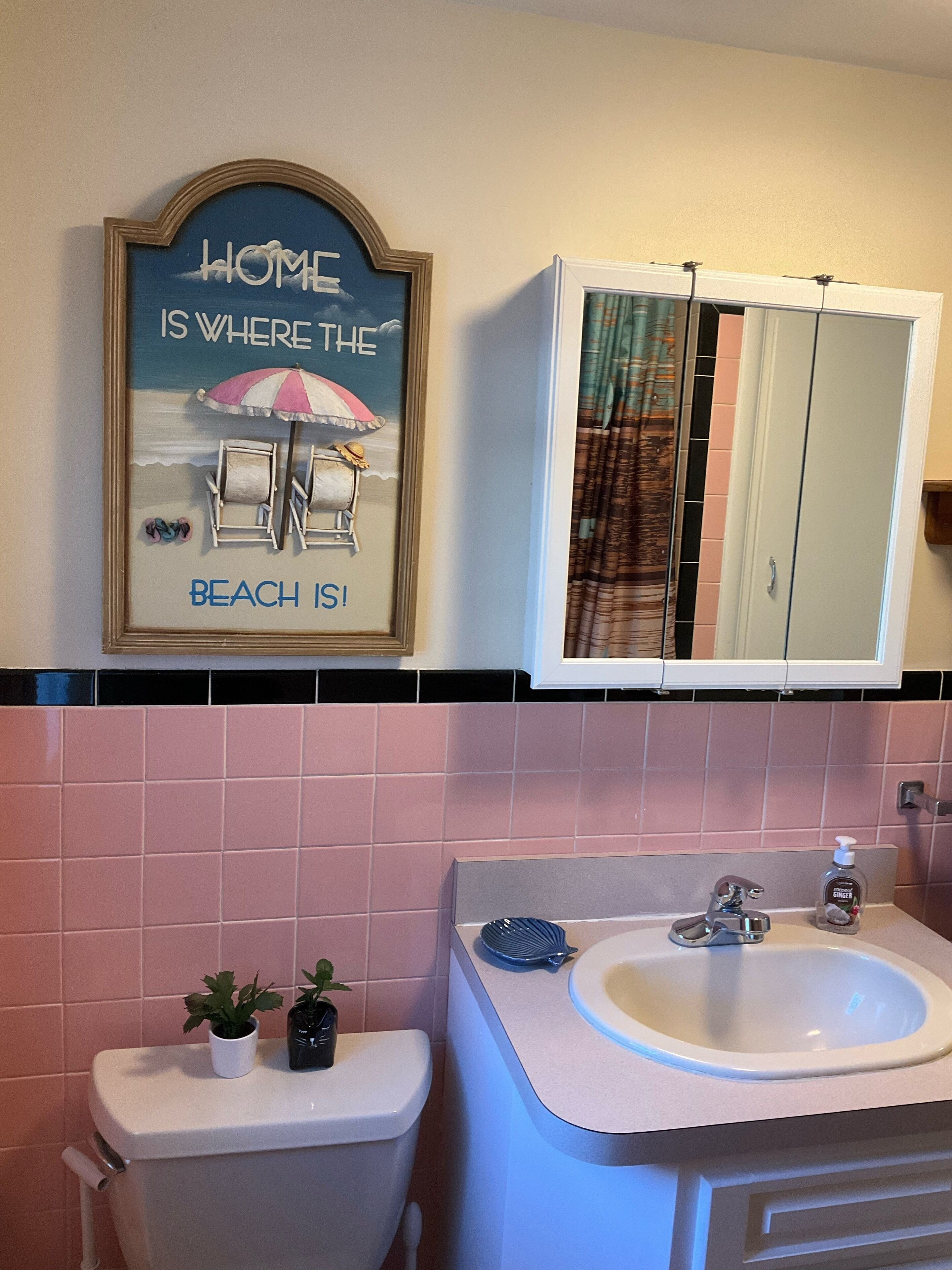 A bathroom with a toilet, sink, and a mirror cabinet. There is a framed picture on the wall above the toilet.