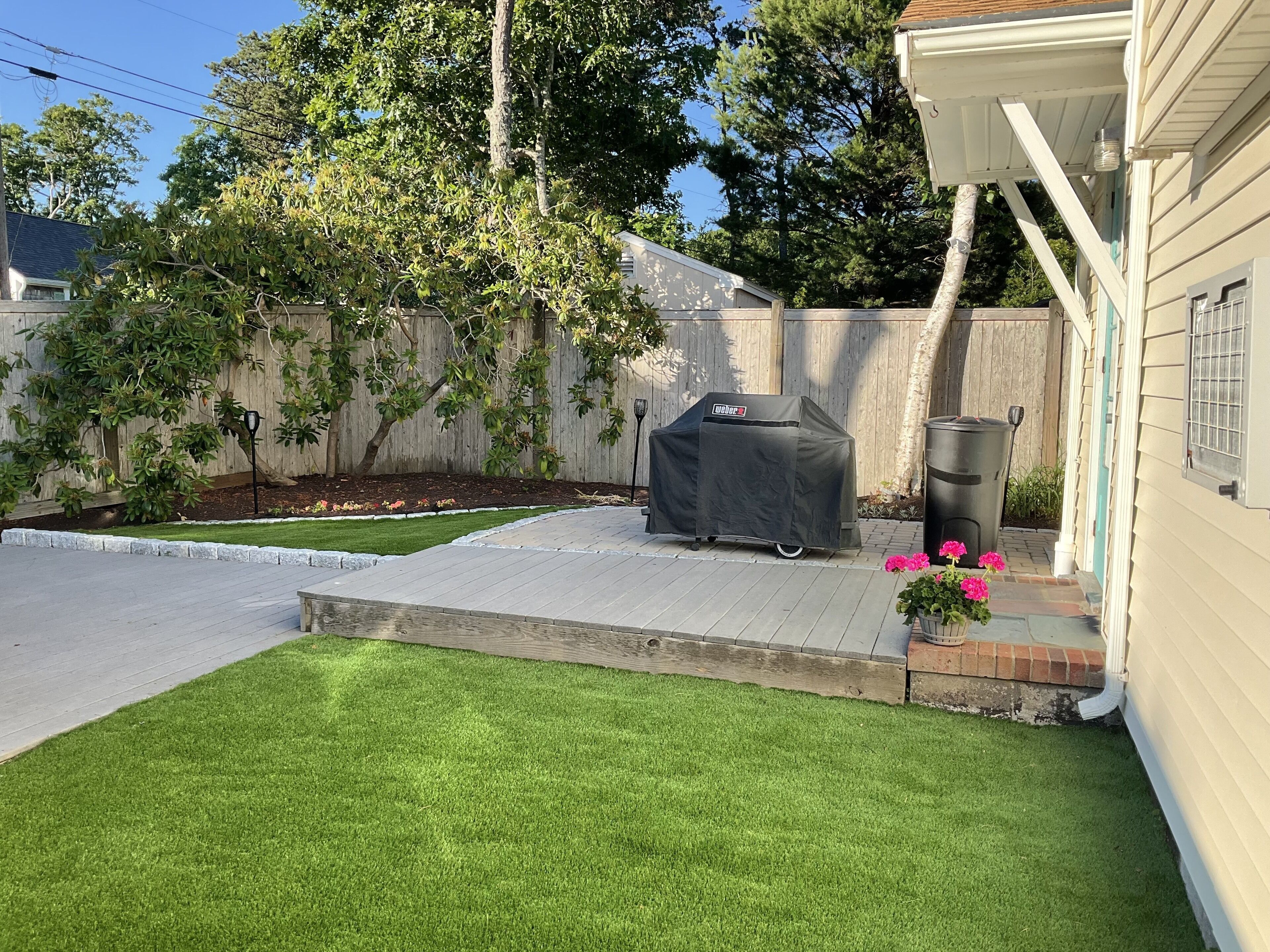 Ein Garten mit Terrasse, Grillhaus, Mülleimer und einem kleinen Garten mit rosa Blumen.