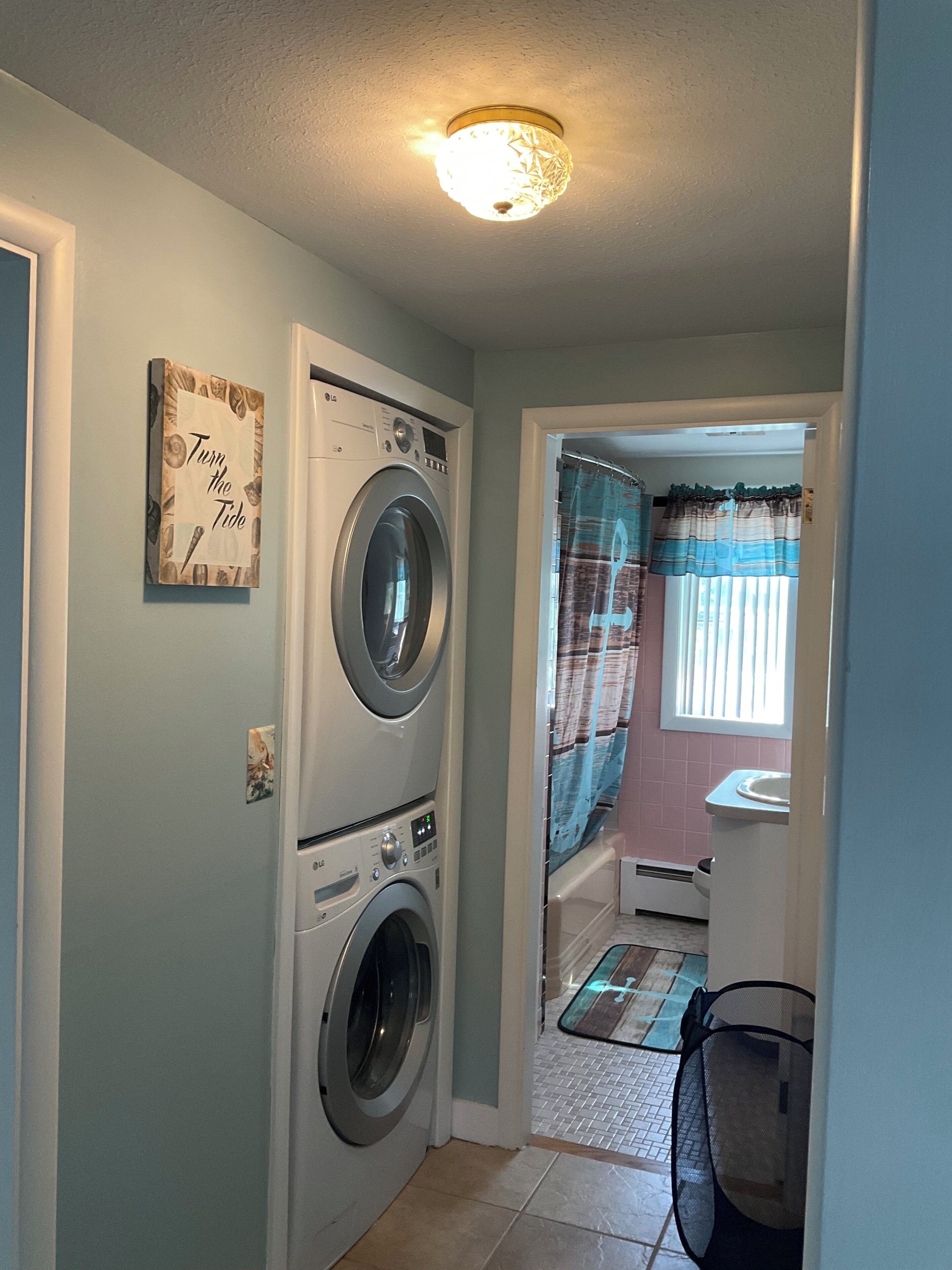 A laundry room with a front-loading washer and dryer, a 'Turn the Tide' sign, and a window with curtains.