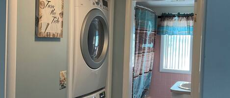 A laundry room with a front-loading washer and dryer, a 'Turn the Tide' sign, and a window with curtains.