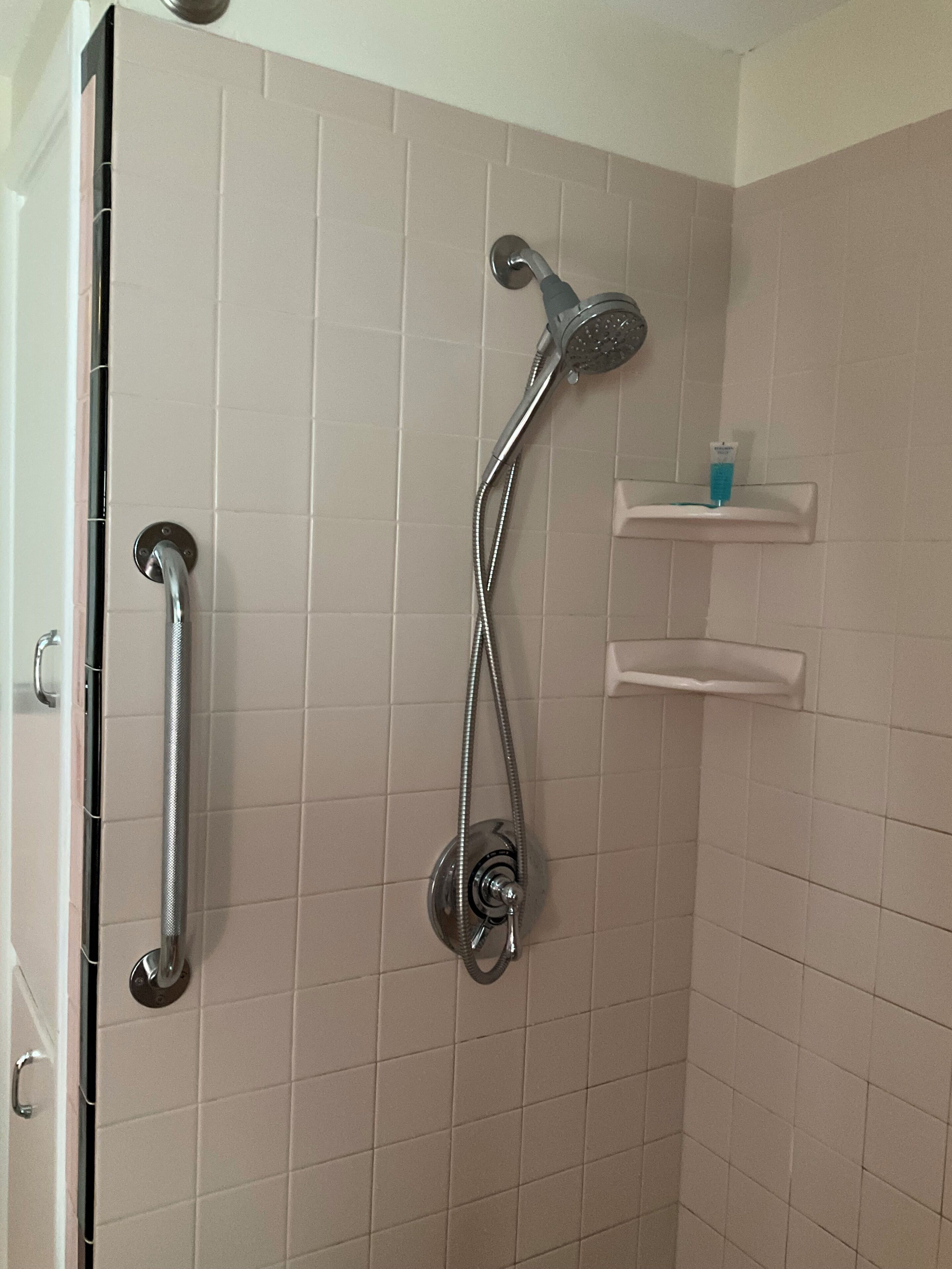 Ducha con cabezal de ducha de mano, grifo empotrado en la pared y un pequeño estante con artículos de aseo.