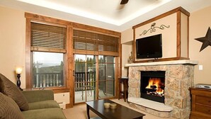 Una sala de estar acogedora con chimenea, un televisor montado encima y un balcón con ventanas amplias.