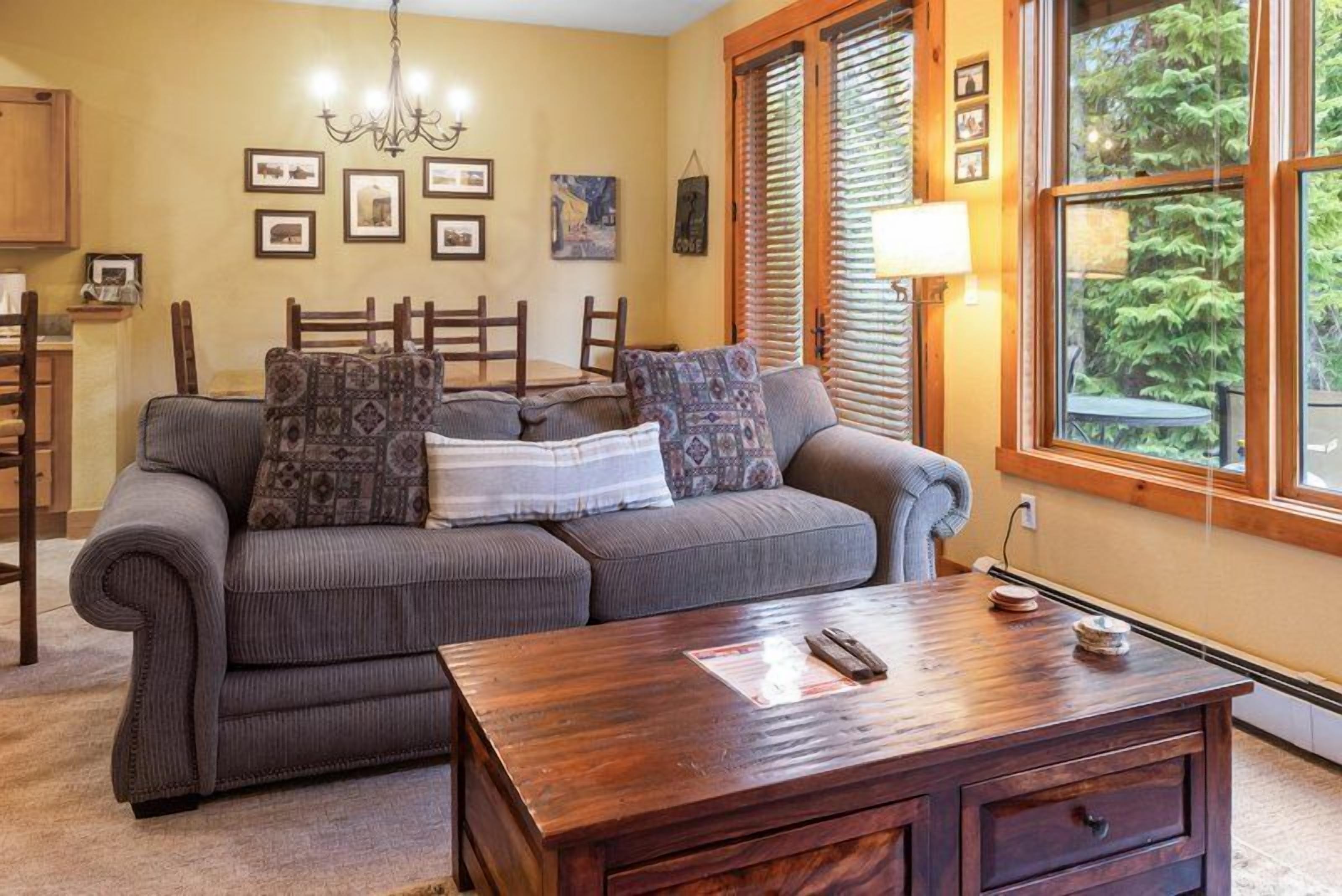 Una sala de estar acogedora con un sofá gris, cojines estampados, una mesa de centro de madera y una ventana con persianas.