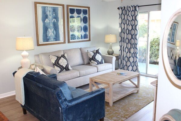 A living room with a sofa, armchair, coffee table, and two lamps. There are two framed artworks on the wall and a patterned curtain. - Pet-friendly Beach Condo, Ciboney 1008 (Destin)