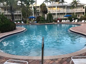 Ein Swimmingpool mit Liegestühlen und Sonnenschirmen, umgeben von Palmen und einem mehrstöckigen Gebäude mit Balkonen.