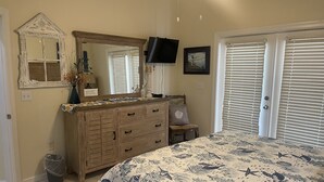 Un dormitorio con cómoda de madera, espejo grande, televisor de pantalla plana, cama con cubrecama de temática marina y ventana con persianas.
