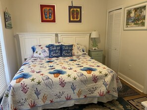 Ein Schlafzimmer mit einem Bett, das mit einer Küsten-Quiltdecke geschmückt ist, zwei gerahmten Kunstwerken über dem Bett, einem Nachttisch mit Lampe und einer Tür mit Blick auf den Strand.
