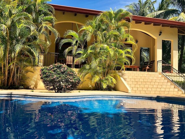 A poolside view of a resort with palm trees, a pool, and a building with a red roof and beige walls.