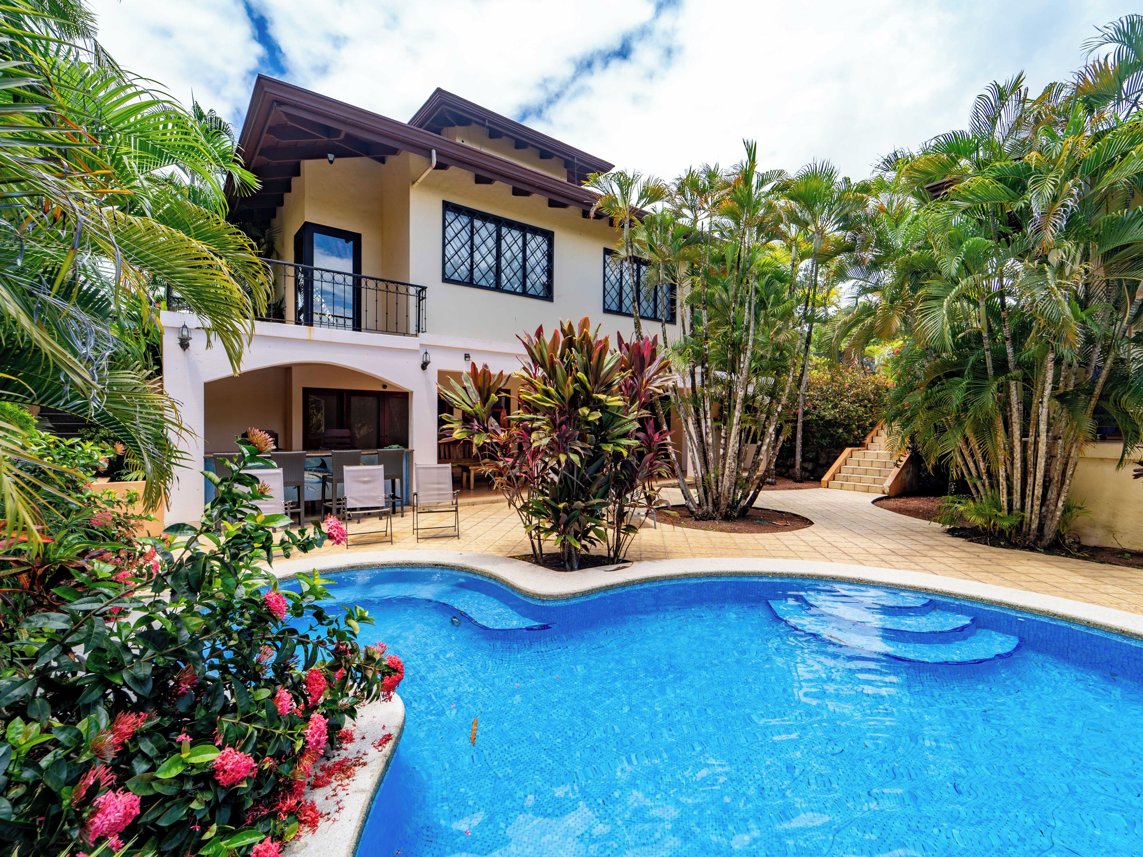 Ein zweistöckiges Haus mit Swimmingpool, umgeben von tropischen Pflanzen und einer Terrassenfläche.