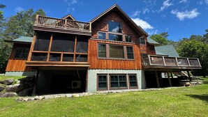 A large wooden house with multiple windows, a balcony, and a garage. -  Sleeps 20! Minutes to Main St & Lakes HotTub PoolTable Pingpong FirePit AC (Lake Placid)
