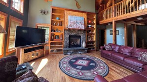 A spacious living room with a fireplace, wooden beams, a television, and a red leather couch. -  Sleeps 20! Minutes to Main St & Lakes HotTub PoolTable Pingpong FirePit AC (Lake Placid)