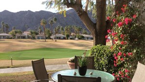 Un patio con una mesa redonda de vidrio y dos sillas, con vista a un campo de golf y montañas al fondo.
