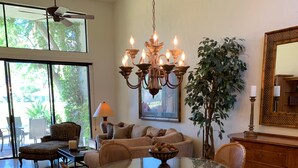Un salón amplio con una mesa redonda de cristal rodeada de sillas de mimbre, un candelabro y una gran planta en maceta.