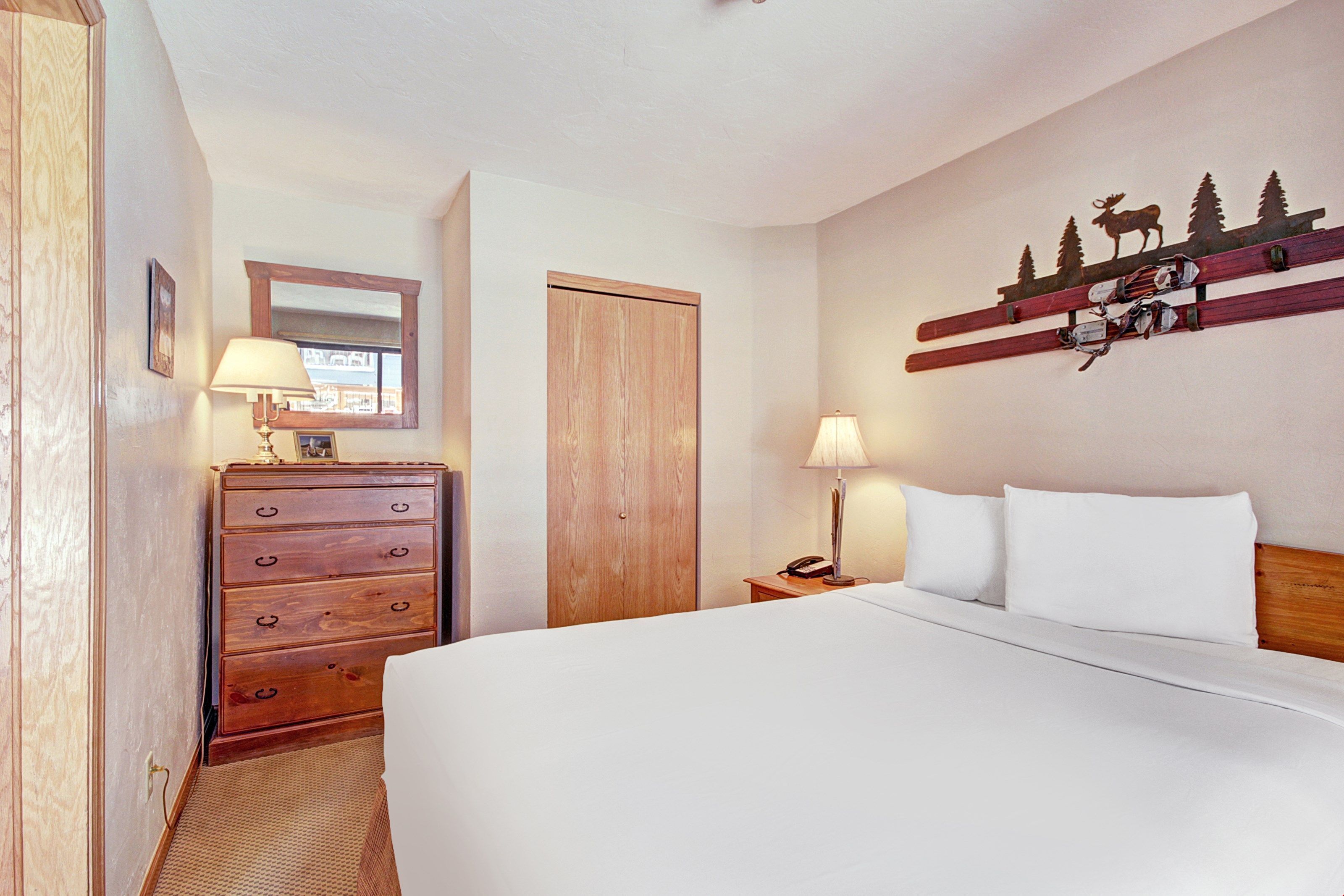 Habitación de hotel con una cama grande, cómoda de madera, lámparas de noche, una puerta de madera y un adorno en la pared con árboles y un alce.