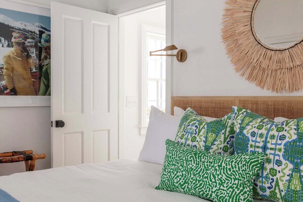 Una cama bien hecha con almohadas estampadas, un espejo colgado en la pared y una puerta que da a otra habitación.