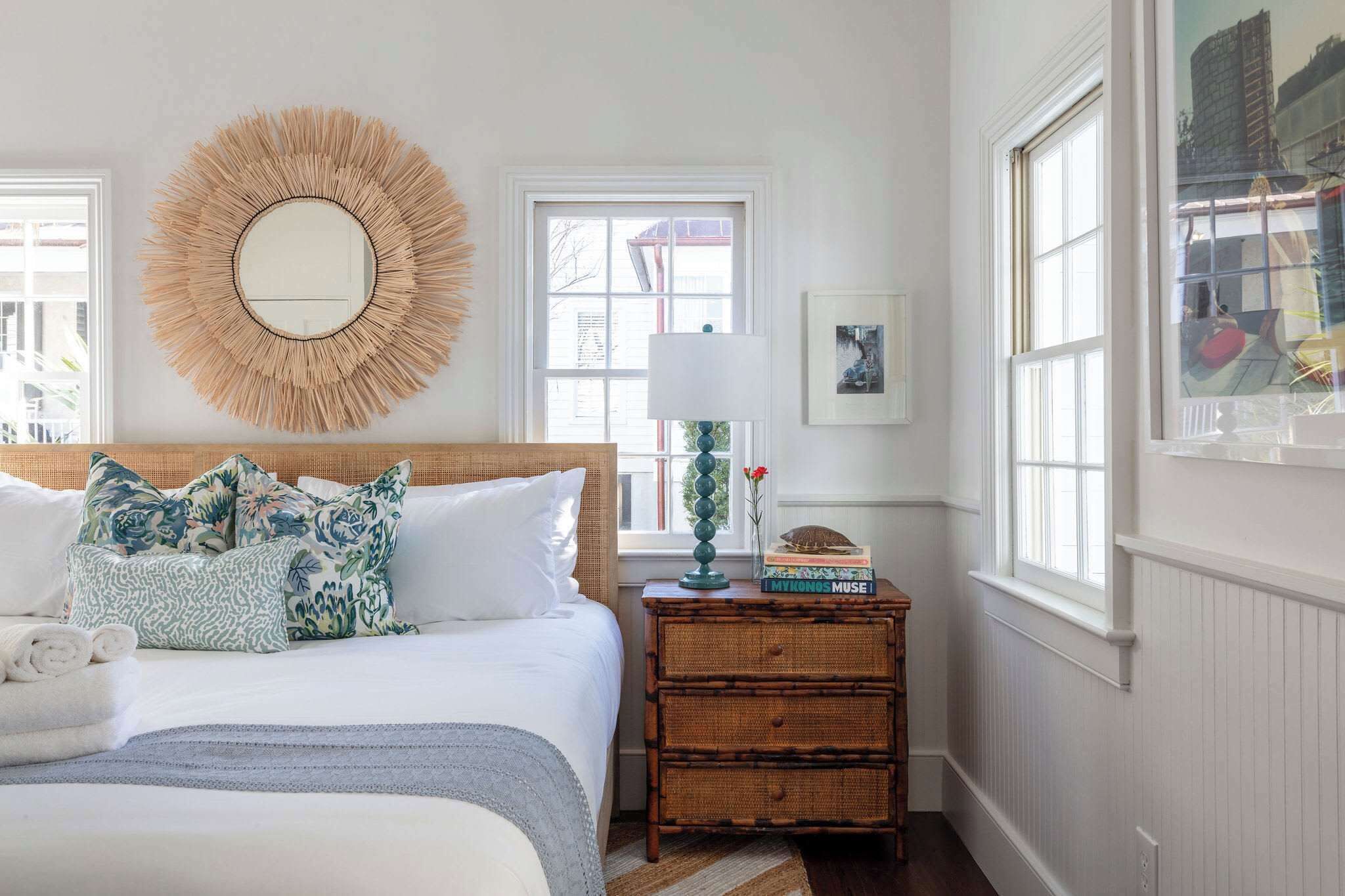 A neatly arranged bedroom with a large bed, a round mirror, a bedside table with a lamp, and a view of the outside through a window.