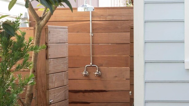 Une cabine de douche extérieure en bois, dotée d’un robinet en métal, entourée d’une clôture en bois et de plantes.