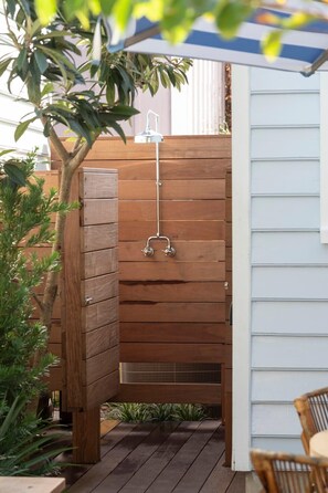 Un box de ducha exterior de madera con un grifo de metal, rodeado por una valla de madera y plantas.
