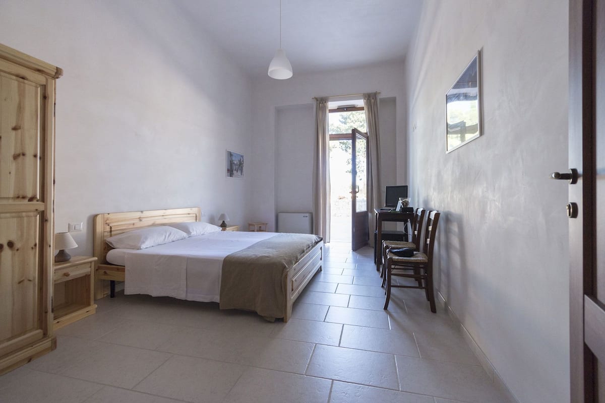A bedroom with a bed, bedside tables, a desk with a chair, a wardrobe, and a door leading to another room.