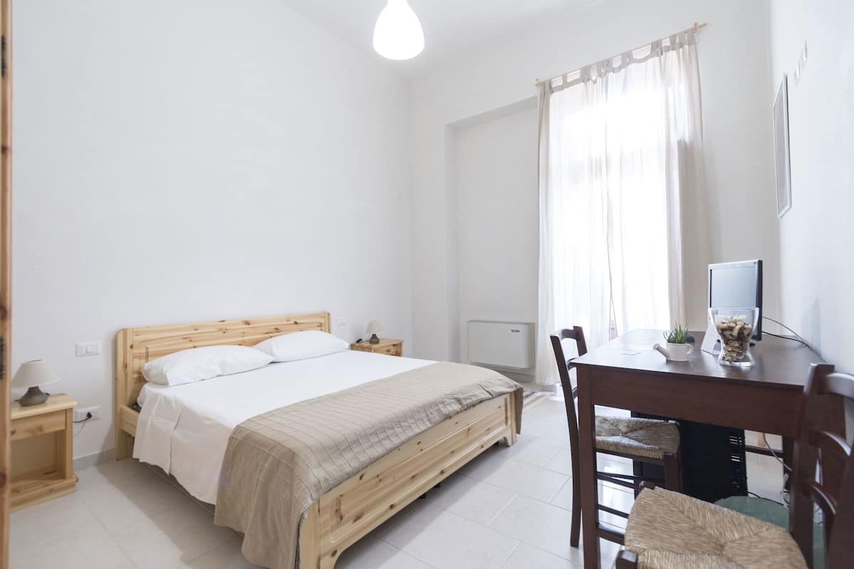 A bedroom with a wooden bed, bedside tables, a desk with a chair, and a window with curtains.