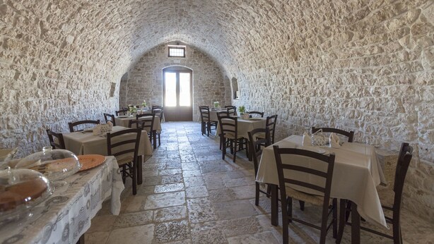 Un comedor con paredes de piedra, mesas y sillas, un techo alto con arcos, y una puerta que da al exterior.