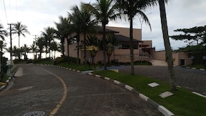 A paved road leading to a building with a modern design, surrounded by palm trees and greenery. - Cond Hanga Roa - Bertioga Riviera (Bertioga)